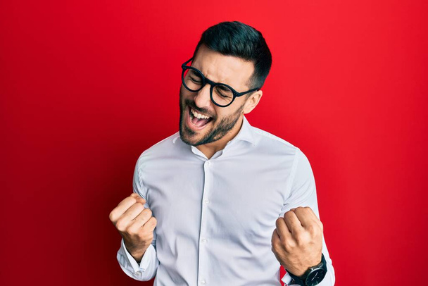 Young hispanic businessman wearing shirt and glasses very happy and excited doing winner gesture with arms raised, smiling and screaming for success. celebration concept.  - 写真・画像
