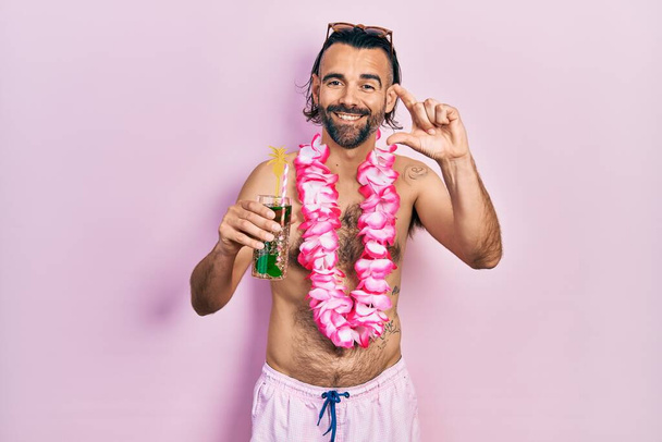 Young hispanic man wearing swimsuit and hawaiian lei drinking tropical cocktail smiling and confident gesturing with hand doing small size sign with fingers looking and the camera. measure concept.  - Photo, Image