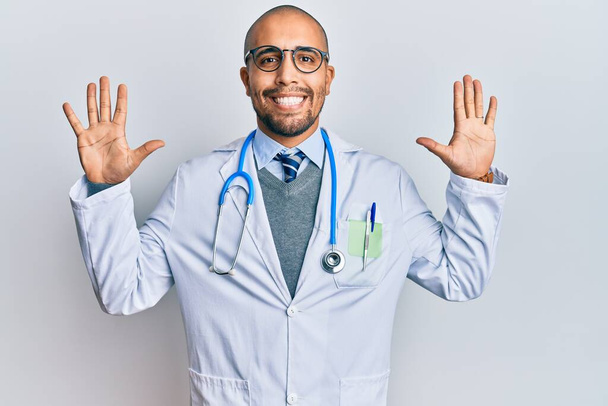 Uomo adulto ispanico indossando uniforme medico e stetoscopio mostrando e indicando con le dita numero dieci, mentre sorridendo fiducioso e felice.  - Foto, immagini