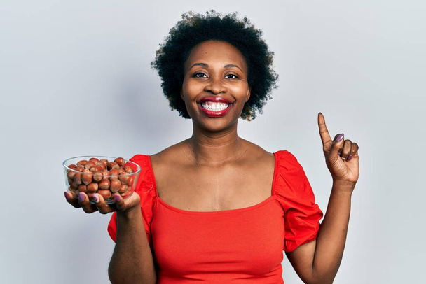 Mujer afroamericana joven sosteniendo avellanas crudas sonriendo con una idea o pregunta señalando el dedo con la cara feliz, número uno  - Foto, Imagen