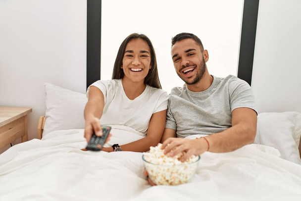 Fiatal latin pár filmet néz és popcornt eszik az ágyon fekve. - Fotó, kép