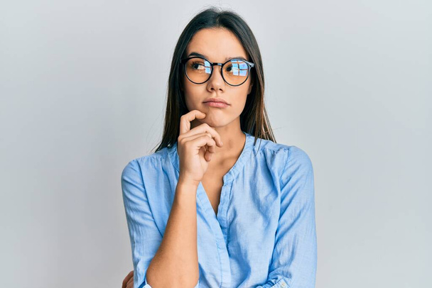 Young hispanic girl wearing casual clothes and glasses thinking concentrated about doubt with finger on chin and looking up wondering  - 写真・画像