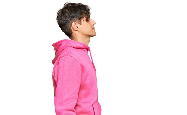 Joven hombre hispano con sudadera rosa casual mirando al costado, pose de perfil relajado con cara natural con sonrisa confiada.  - Foto, Imagen