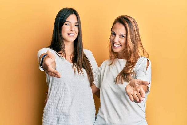 Ισπανόφωνη οικογένεια της μητέρας και της κόρης φορώντας casual λευκό μπλουζάκι χαμογελώντας χαρούμενα προσφέροντας παλάμη χέρι δίνοντας βοήθεια και αποδοχή.  - Φωτογραφία, εικόνα