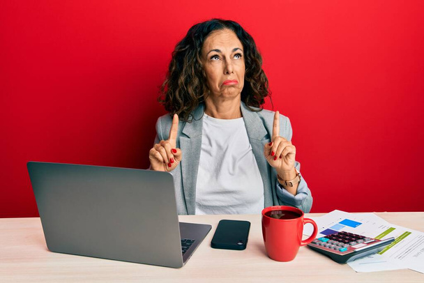 Ofiste çalışan orta yaşlı güzel bir kadın bir fincan kahve içiyor üzgün ve üzgün görünüyor, parmaklarıyla yönü gösteriyor, mutsuz ve depresif..  - Fotoğraf, Görsel