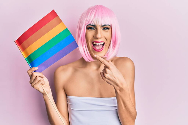 Joven hombre usando mujer maquillaje sosteniendo arco iris lgbtq bandera con peluca rosa sonriendo feliz señalando con la mano y el dedo  - Foto, imagen