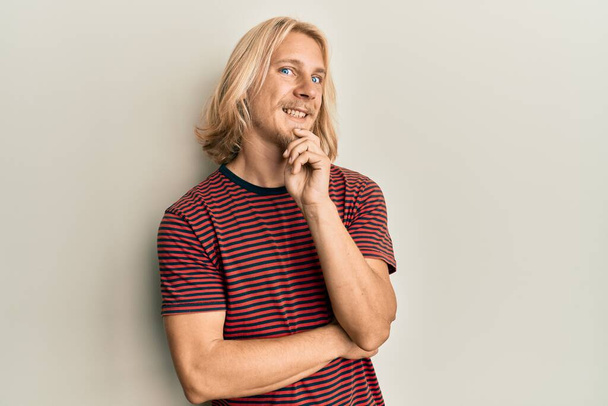 Caucasian young man with long hair wearing casual striped t shirt smiling looking confident at the camera with crossed arms and hand on chin. thinking positive.  - Foto, immagini
