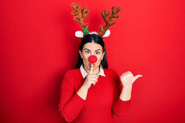 Молода іспаномовна жінка носить різдвяний капелюх оленя і червоний ніс з проханням мовчати з пальцем на губах, спрямованим рукою в сторону. тиша і таємна концепція
.  - Фото, зображення