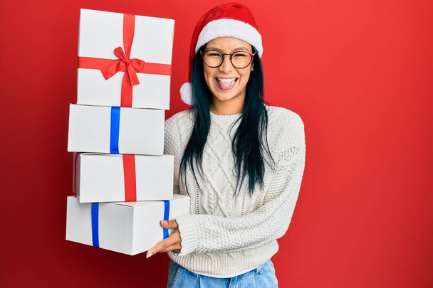 Beautiful hispanic woman wearing christmas hat and holding gifts sticking tongue out happy with funny expression.  - Photo, Image