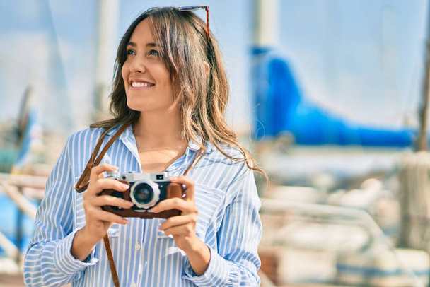 Молодая испаноязычная туристка улыбается счастливой, используя винтажную камеру в порту. - Фото, изображение