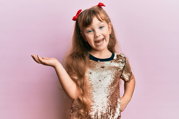 小さなcaucasian女の子子供身に着けているお祝いのスパンコールドレス笑顔陽気なプレゼンテーションとポインティングとともに手のひらの手見てカメラ.  - 写真・画像