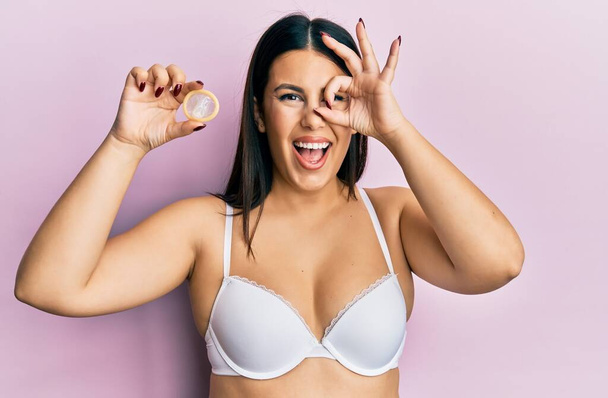 Beautiful brunette woman wearing lingerie and holding condom smiling happy doing ok sign with hand on eye looking through fingers  - Фото, изображение
