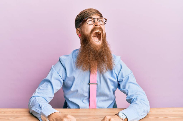 Молодой ирландский рыжий мужчина в деловой рубашке и галстуке сидит на столе злой и безумный крича расстроенный и разъяренный, крича от злости. гнев и агрессивная концепция.  - Фото, изображение