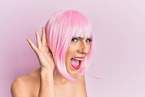Молодий чоловік у жінці одягнений у рожеву перуку, що посміхається рукою через вухо, слухаючи чутки чи плітки. концепція глухоти
.  - Фото, зображення