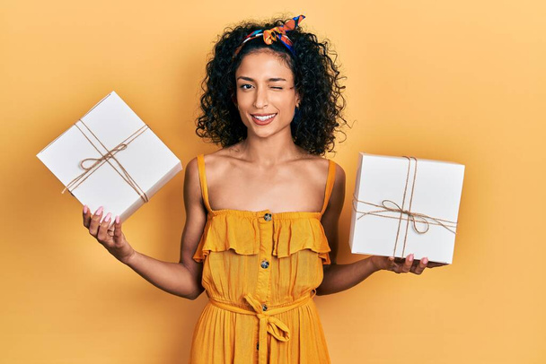 Joven chica latina sosteniendo regalos guiñando el ojo mirando a la cámara con expresión sexy, cara alegre y feliz.  - Foto, imagen
