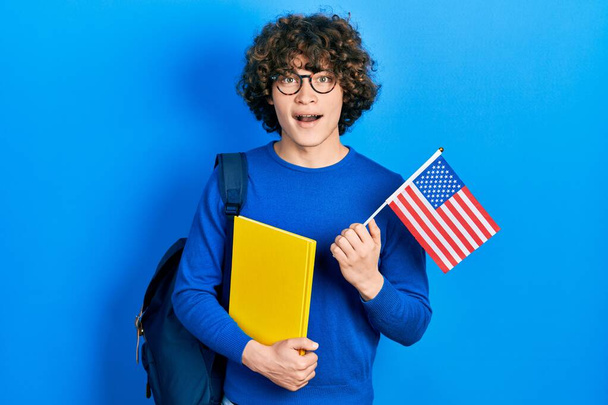 Beau jeune homme échange étudiant tenant le drapeau des Etats-Unis célébrant fou et étonné pour le succès avec les yeux ouverts hurlant excité.  - Photo, image