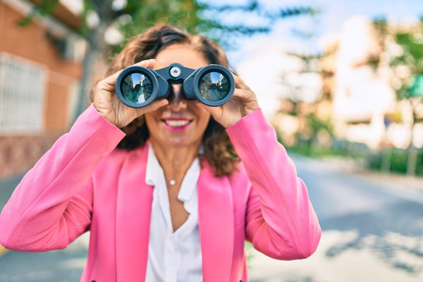 Empresaria hispana de mediana edad sonriendo feliz buscando una nueva oportunidad usando prismáticos en la ciudad. - Foto, Imagen