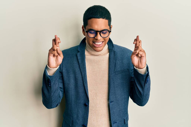 Young handsome hispanic man wearing elegant clothes and glasses gesturing finger crossed smiling with hope and eyes closed. luck and superstitious concept.  - Photo, image