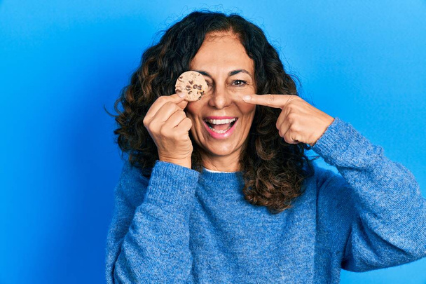 Middelbare leeftijd Spaanse vrouw houdt koekje over oog glimlachend gelukkig wijzend met hand en vinger  - Foto, afbeelding