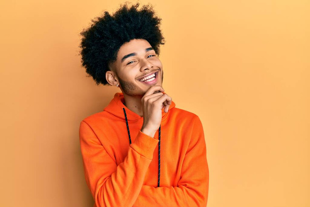 Young african american man with afro hair wearing casual sweatshirt smiling looking confident at the camera with crossed arms and hand on chin. thinking positive.  - Foto, Imagem