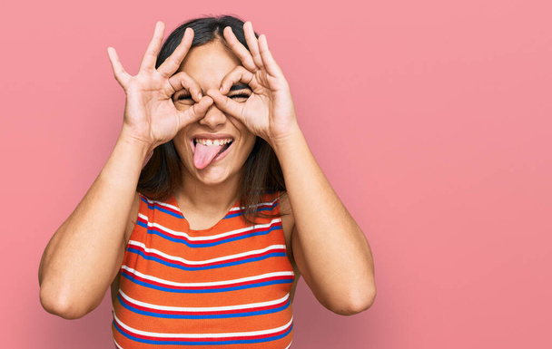 Young brunette woman wearing casual clothes doing ok gesture like binoculars sticking tongue out, eyes looking through fingers. crazy expression.  - Foto, Bild