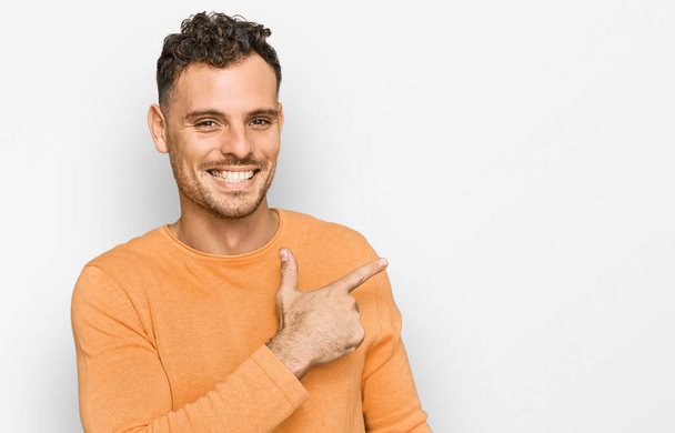 Νεαρός ισπανόφωνος άνδρας φορώντας casual ρούχα χαρούμενος με χαμόγελο στο πρόσωπο δείχνοντας με το χέρι και το δάχτυλο στο πλάι με χαρούμενη και φυσική έκφραση στο πρόσωπο  - Φωτογραφία, εικόνα