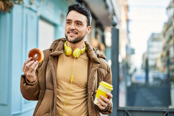 Young hispanic man having breakfast using headphones at the city. - Photo, Image