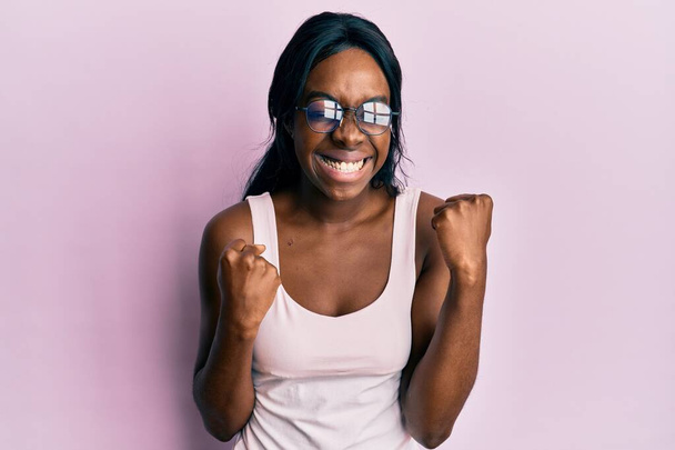 Молода афроамериканська жінка, одягнена в повсякденний одяг і окуляри, які відзначали здивований і вражений успіхом з піднятими руками і закритими очима  - Фото, зображення