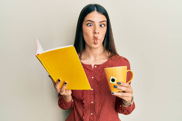 Young hispanic woman reading book drinking cup of tea making fish face with mouth and squinting eyes, crazy and comical.  - Photo, Image