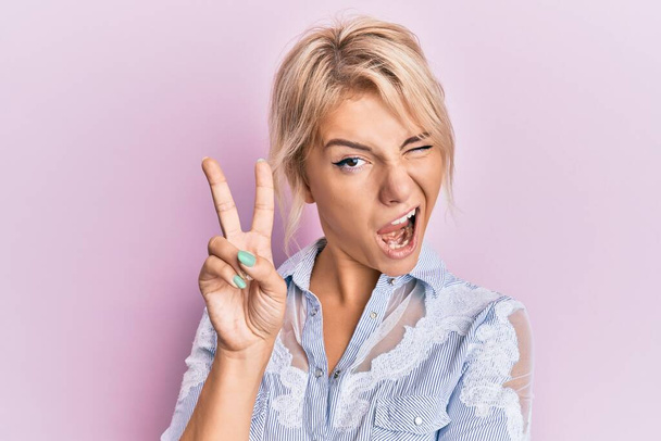 Young blonde girl wearing casual clothes smiling with happy face winking at the camera doing victory sign. number two.  - Photo, Image