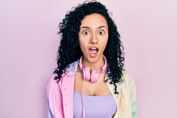 Young hispanic woman with curly hair wearing gym clothes and using headphones afraid and shocked with surprise expression, fear and excited face.  - Photo, Image