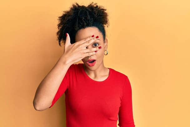 Νεαρή Αφρο-Αμερικανίδα που φοράει καθημερινά ρούχα κρυφοκοιτάζει σε κατάσταση σοκ καλύπτοντας το πρόσωπο και τα μάτια με το χέρι, κοιτάζοντας μέσα από τα δάχτυλα φοβούμενη  - Φωτογραφία, εικόνα