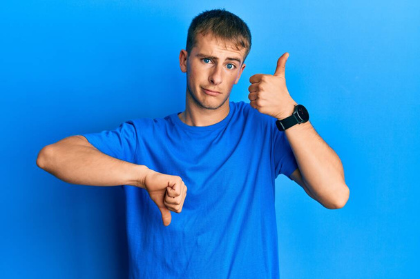 Молодой кавказский мужчина в обычной синей футболке делает большие пальцы вверх и вниз, разногласия и выражение согласия. сумасшедший конфликт  - Фото, изображение