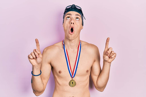 Young hispanic man wearing swimmer glasses and gold medal amazed and surprised looking up and pointing with fingers and raised arms.  - Zdjęcie, obraz