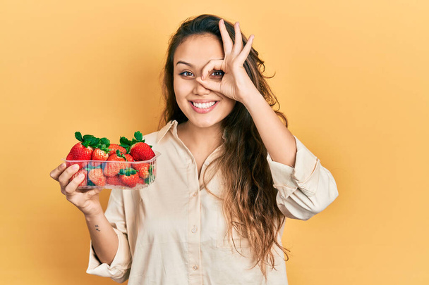 Joven chica hispana sosteniendo fresas haciendo buen gesto con la mano sonriente, los ojos mirando a través de los dedos con cara feliz.  - Foto, Imagen