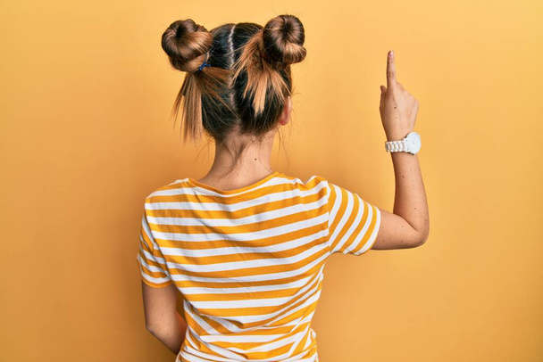 Beautiful brunette little girl wearing casual striped t shirt posing backwards pointing ahead with finger hand  - Photo, Image
