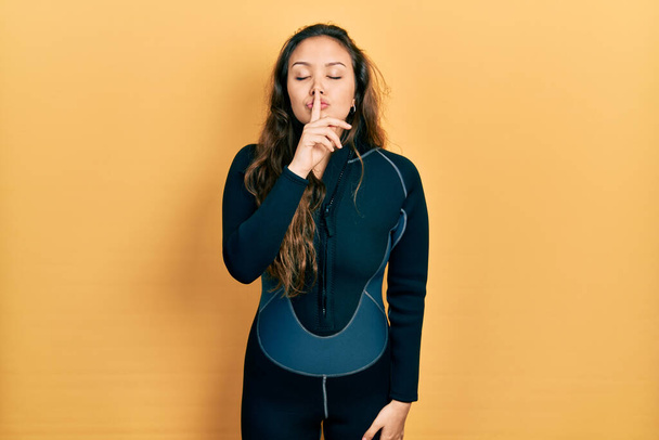 Menina hispânica jovem vestindo uniforme de neoprene mergulhador pedindo para ficar quieto com o dedo nos lábios. silêncio e conceito secreto.  - Foto, Imagem
