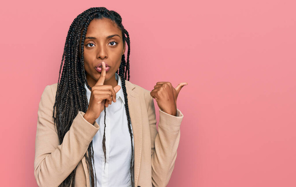 Mujer afroamericana con chaqueta de negocios pidiendo silencio con el dedo en los labios apuntando con la mano hacia un lado. silencio y concepto secreto.  - Foto, imagen