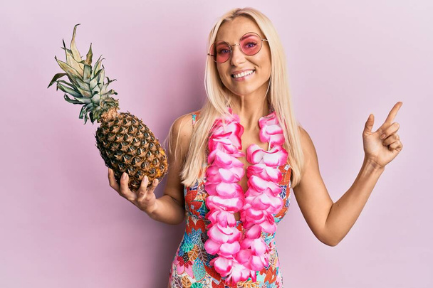 Junge blonde Frau im Badeanzug und hawaiianischem Lei, Ananas lächelnd glücklich mit Hand und Finger zur Seite zeigend  - Foto, Bild