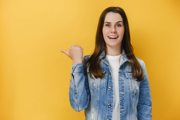 Positive excited young brunette woman 20s points thumb, demonstrates copy space on blank yellow studio background wall, has happy friendly expression, wears blue denim jacket. People emotion concept - Valokuva, kuva