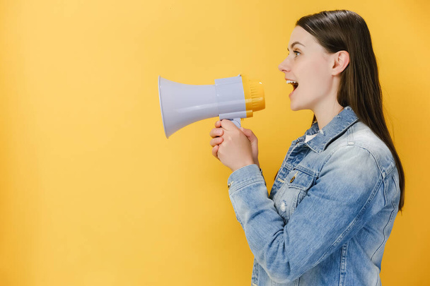Side view of cheerful young woman 20s screaming in megaphone turn around camera, wears denim jacket, posing isolated on yellow studio background wall with copy space. People emotion lifestyle concept - 写真・画像