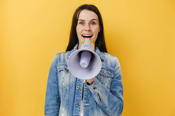 Portrait of fun cute young brunette woman scream in megaphone announces discounts sale hot news Hurry up, wears denim jacket, isolated on yellow studio background. People emotion lifestyle concept - Fotografie, Obrázek