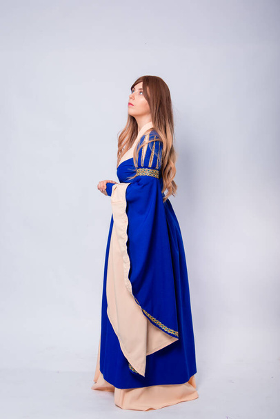 A full-length portrait of an attractive woman with long hair in a medieval, fantasy, blue and beige dress with long, large sleeves, posing isolated on a white background. - Фото, зображення