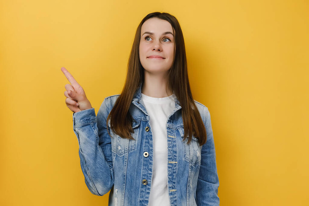 Adorable smiling young woman 20s has positive expression, indicates at blank copy space, shows direction, wears denim jacket, stands against yellow studio background wall. People emotion concept - Fotografie, Obrázek