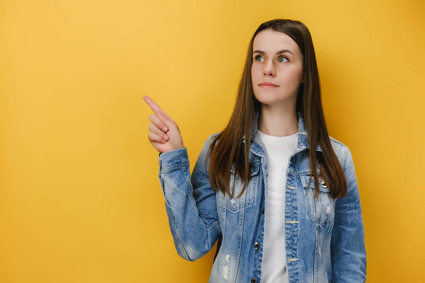 Serious looking caucasian woman indicates away, convinces customer to buy her product, wears denim jacket and white t-shirt, has thoughtful look, isolated on yellow background. Advertising concept - Photo, Image