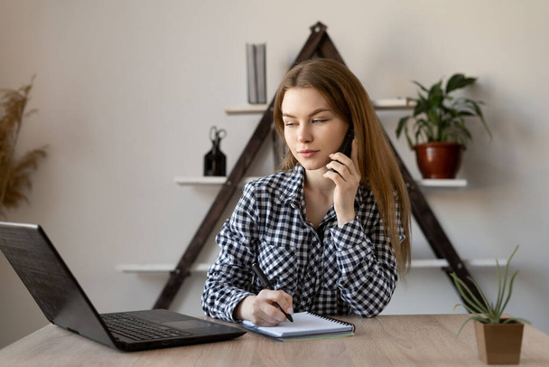 Young business woman talking on the phone negotiates with a client while sitting in the office at a table with a laptop. Smartphone and laptop in the work of an office worker or teacher. - 写真・画像
