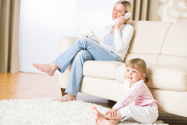 Mother and child in living room watch television, mother on phone in background on sofa - Foto, Bild