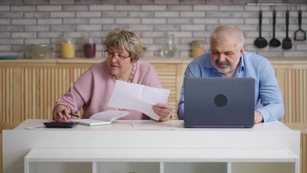 starší manželé zkoumají účty za elektřinu, manželka a manžel si prohlíží papír, počítají s kalkulačkou a platí online notebookem - Záběry, video