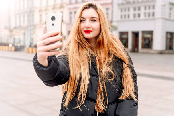 Beautiful girl takes selfie photo. Blonde with phone on city street. Young woman posing for social networks - Photo, Image