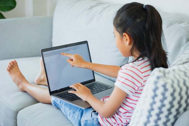 Азійська дівчинка сидить на дивані і робить домашнє завдання в комп'ютерному ноутбуку. Дитяче домашнє навчання під час карантину через спалах COVID-19. - Фото, зображення